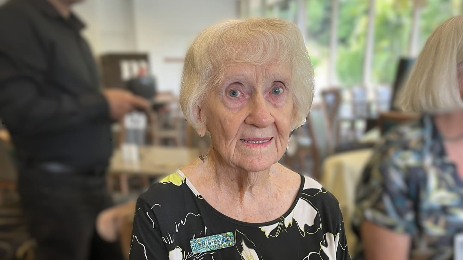 St George Community client Judy, aged 90, sitting at a café table holding a pink drink and smiling during a social outing.